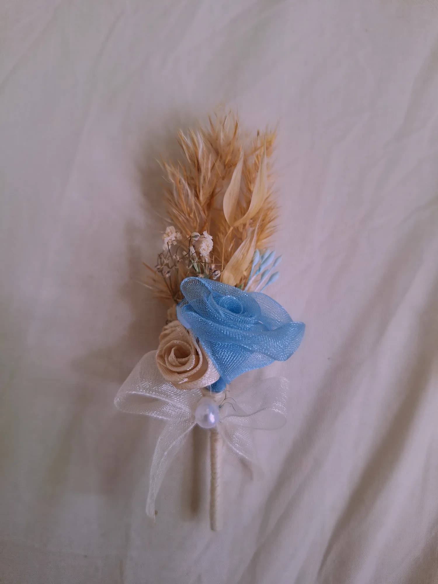 Boutonniere with a pale blue organza rose, a champagne satin rose, dried pampas, oats and gypsophila with an ivory bow and a pearl.